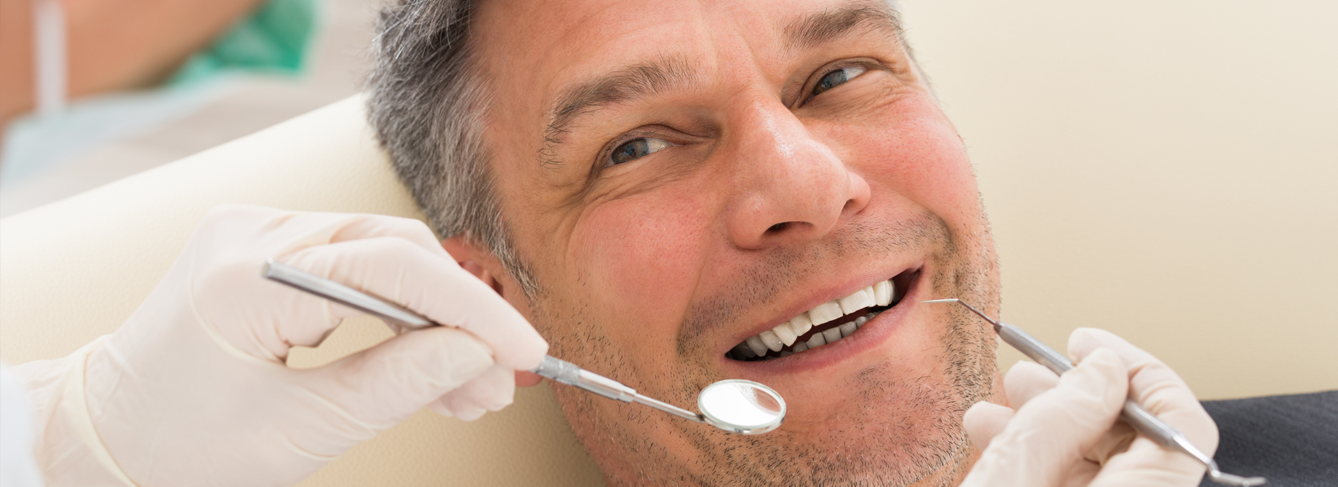 A man with a toothy smile is seated in a dental chair, receiving a dental procedure by a dentist wearing white gloves and using dental tools.